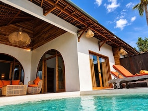 Un área junto a la piscina con mobiliario de mimbre, techo de madera y cielo azul despejado.