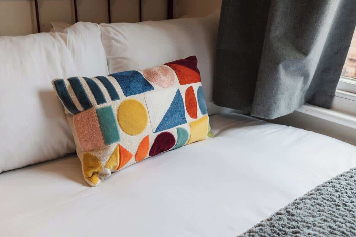 A bed with a pillow featuring a geometric pattern, a white bedspread, and a window with a curtain.