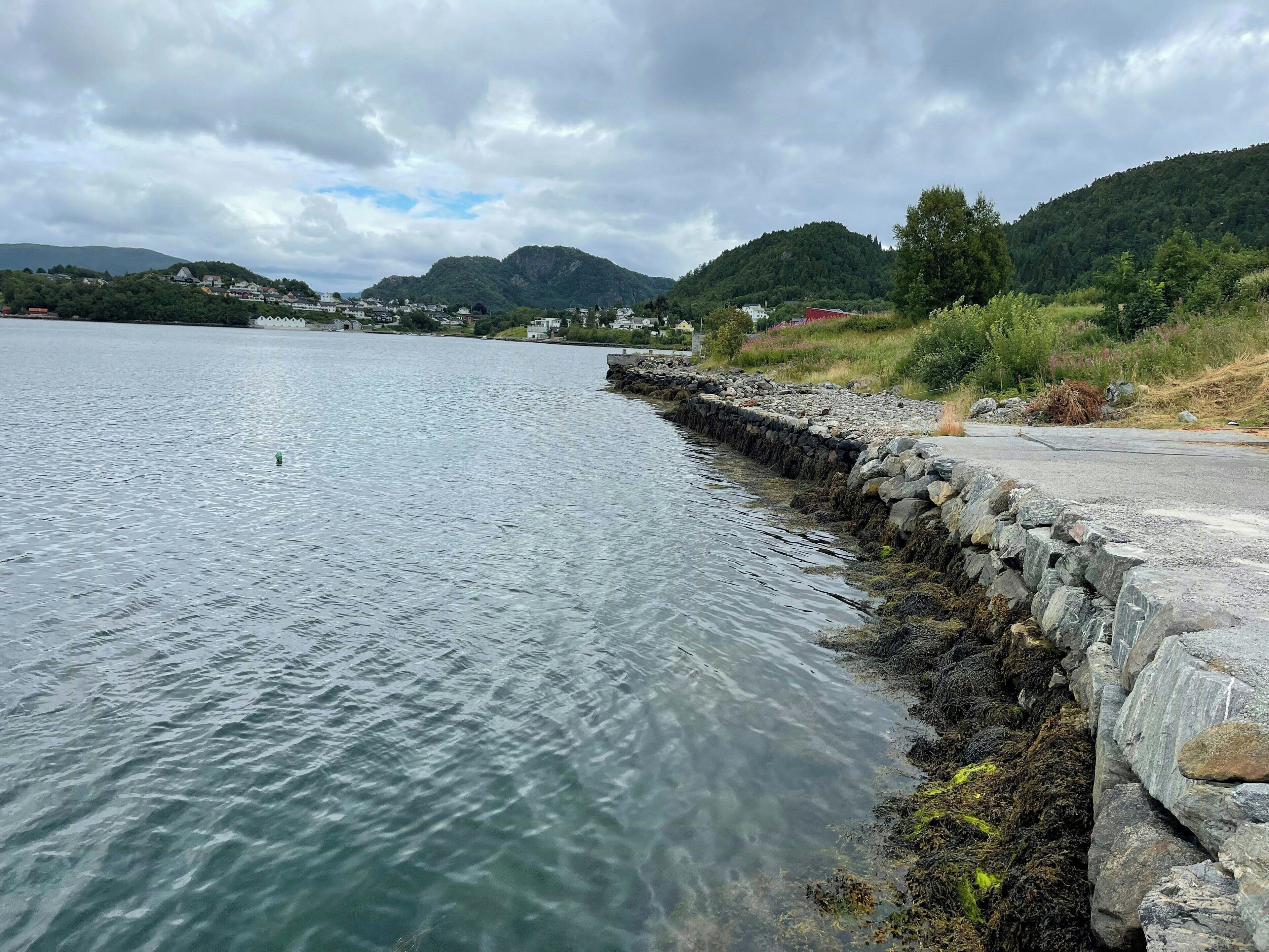 Une vue sur le bord de l’eau, avec un mur de pierre, une eau calme et des collines lointaines.