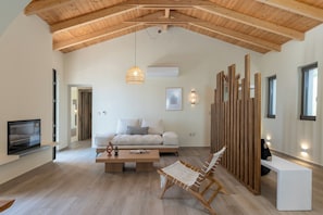 A spacious living room with a high wooden ceiling, a white sofa, a wooden coffee table, a wicker chair, and a TV mounted on the wall. - Orion Sunset Villa (Erissos)