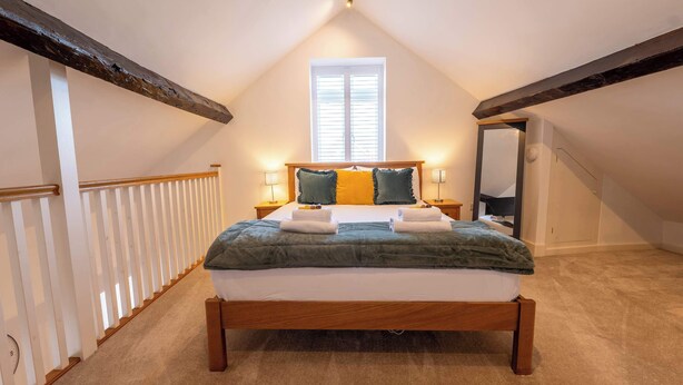 Una camera da letto con soffitto spiovente, un letto con struttura in legno, due lampade sui comodini, una finestra con persiane e una ringhiera bianca.