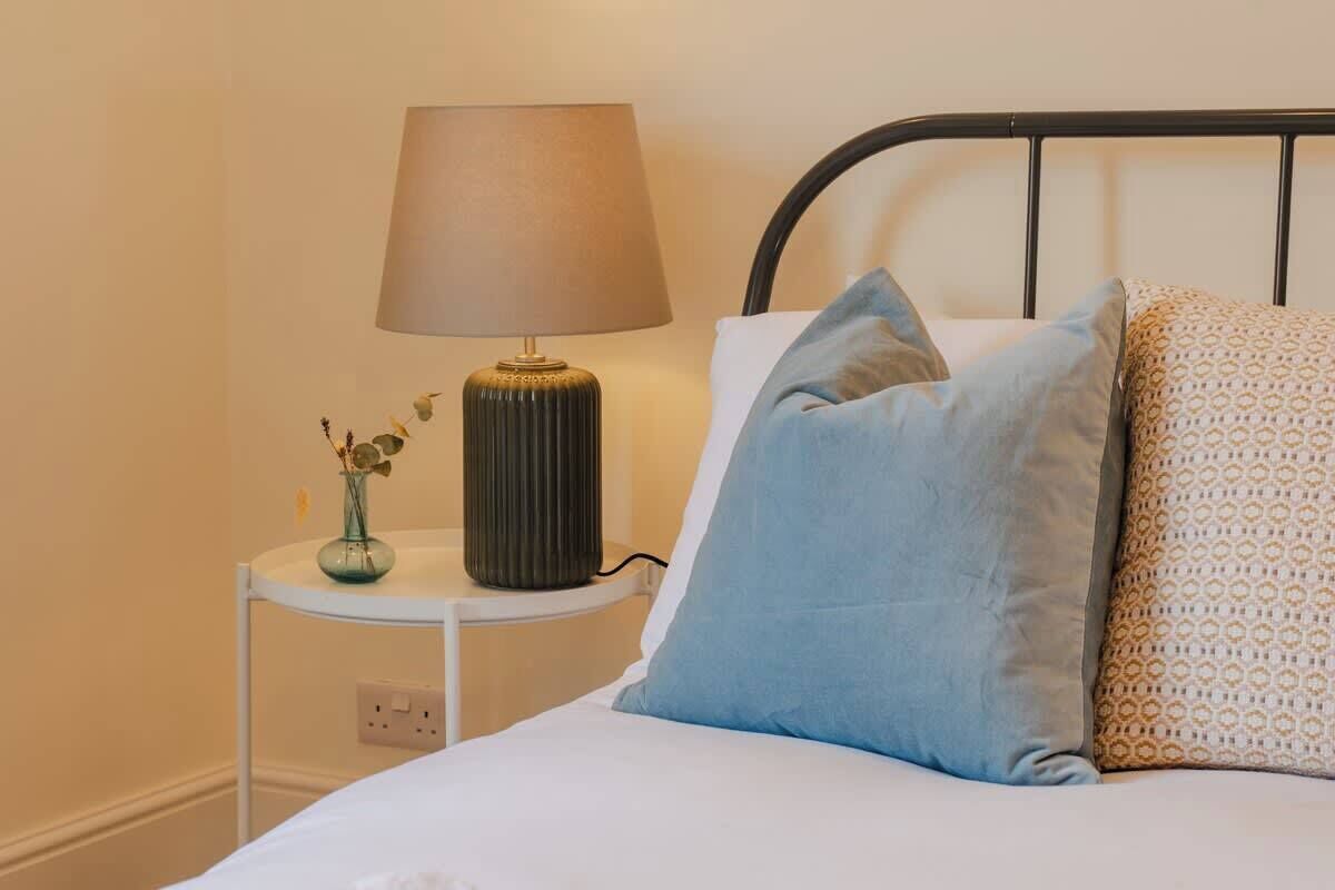 Ein Bett mit weißer Bettdecke, einem blauen Kissen und einem gemusterten Kissen, eine Nachttischlampe und eine kleine Vase mit Blumen auf einem Nachttisch.