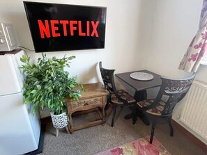 Et værelse med et fladskærms-tv, der viser 'NETFLIX', et træbord med stole, en potteplante og en radiator.