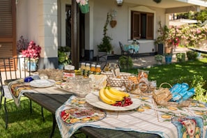 A table set outdoors with a tablecloth, plates, glasses, a basket of bread, and a fruit display.