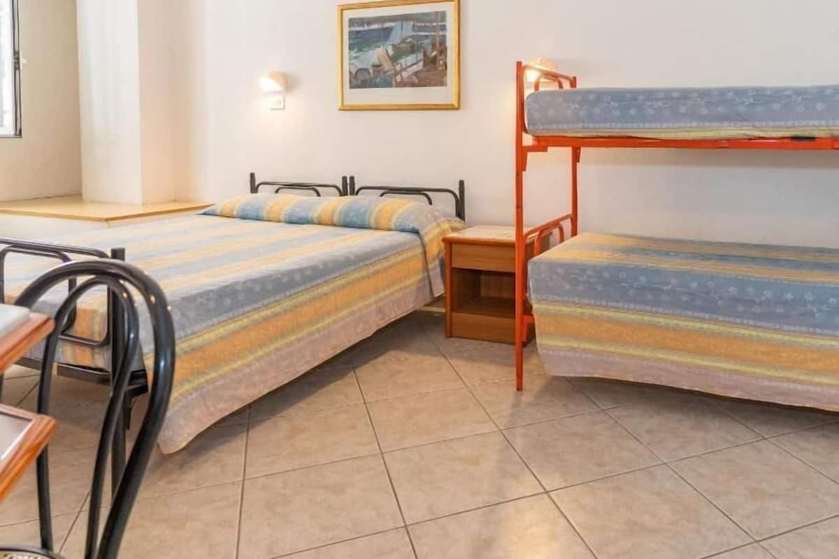 A dormitory room with two beds, a desk, a chair, a small table, and a wall-mounted light.