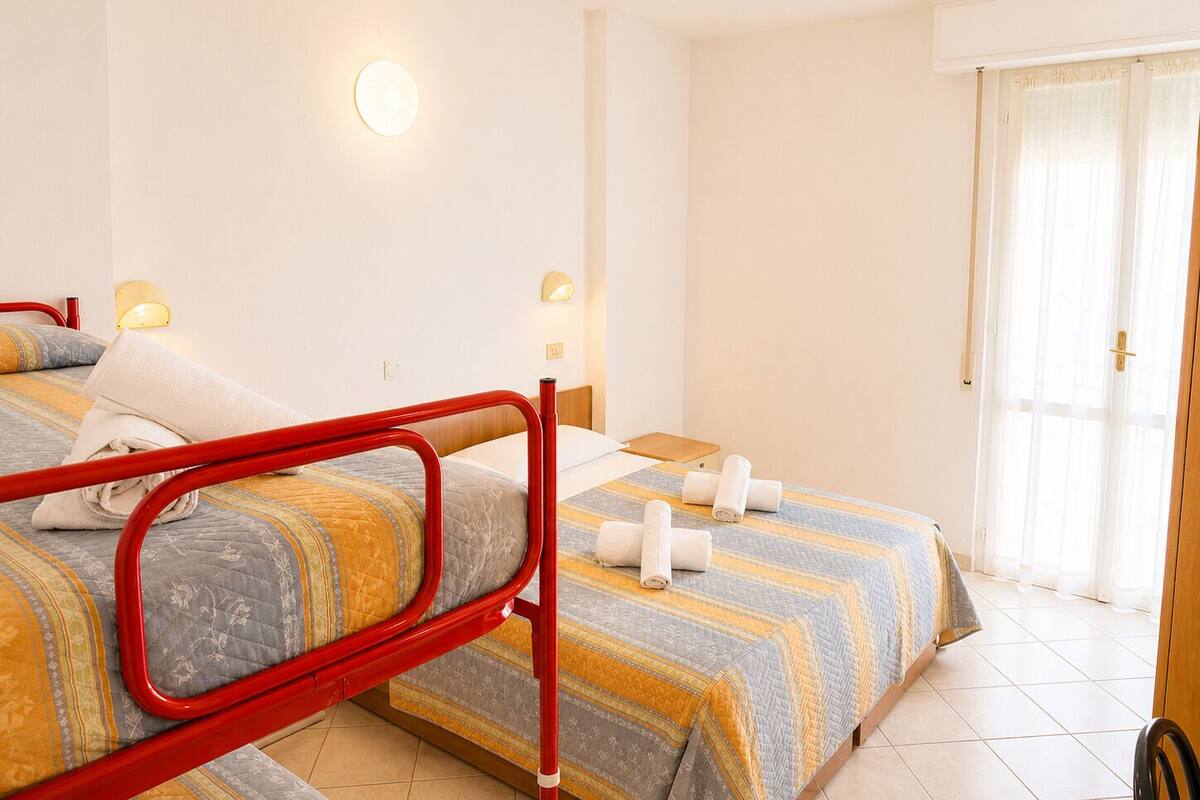 A twin room with two beds, a red metal bed frame, two bedside tables, a wall-mounted light, and a sliding glass door.