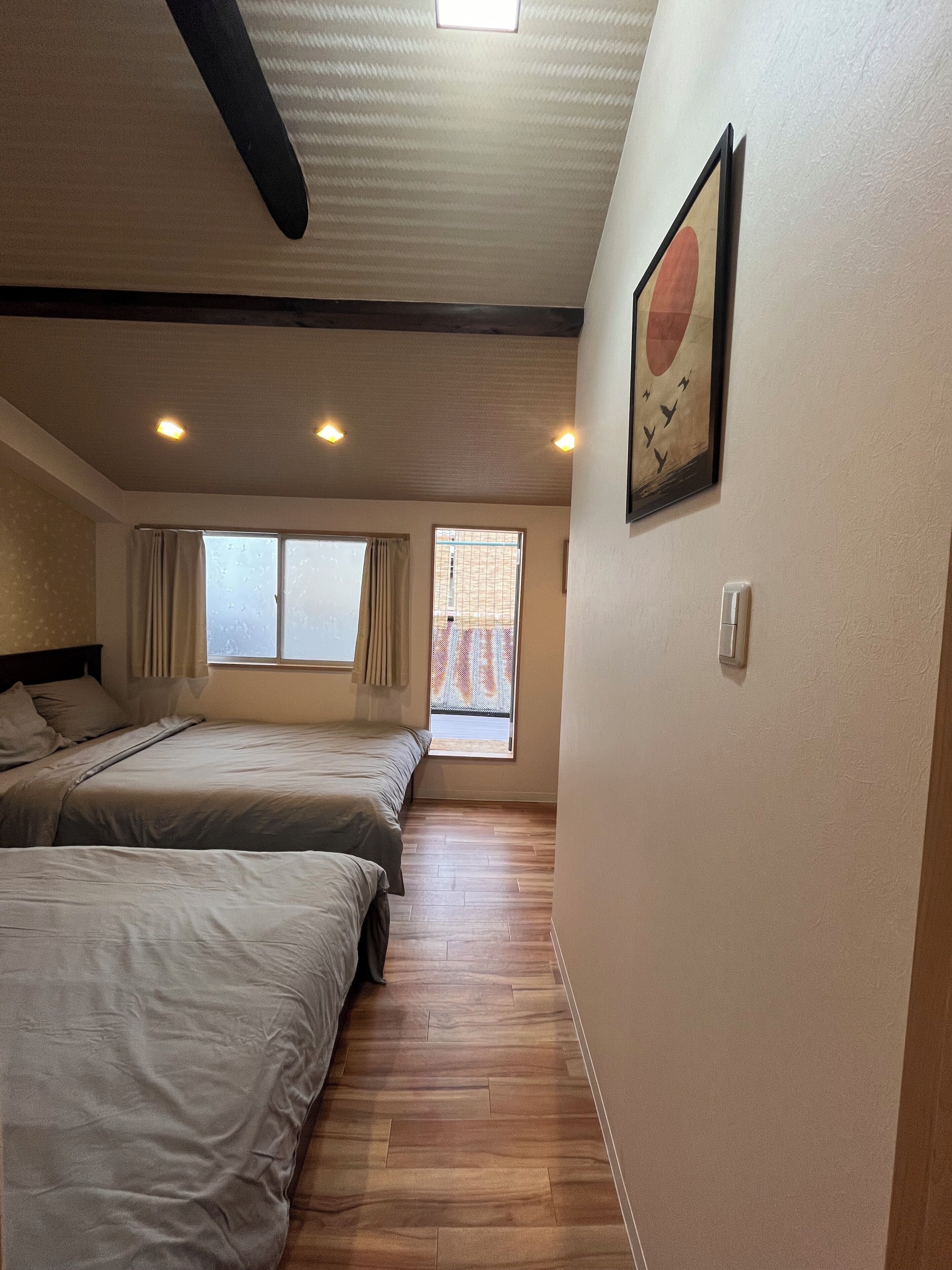 Habitación de hotel con dos camas, una ventana con cortinas, suelo de madera, techo con iluminación empotrada e imagen colgada en la pared.