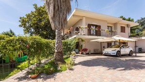 Una casa a due piani con un'auto nel vialetto, circondata da piante in vaso e un albero.