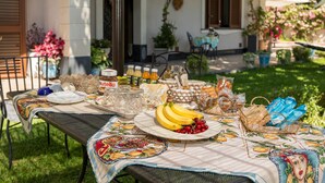 Un tavolo apparecchiato all'aperto con tovaglia, piatti, bicchieri, un cestino di pane e un cesto di frutta.