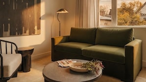 A modern living room with a green sofa, a round coffee table with magazines and a vase, a floor lamp, and a view of trees through a window. - Ceros (Denver)