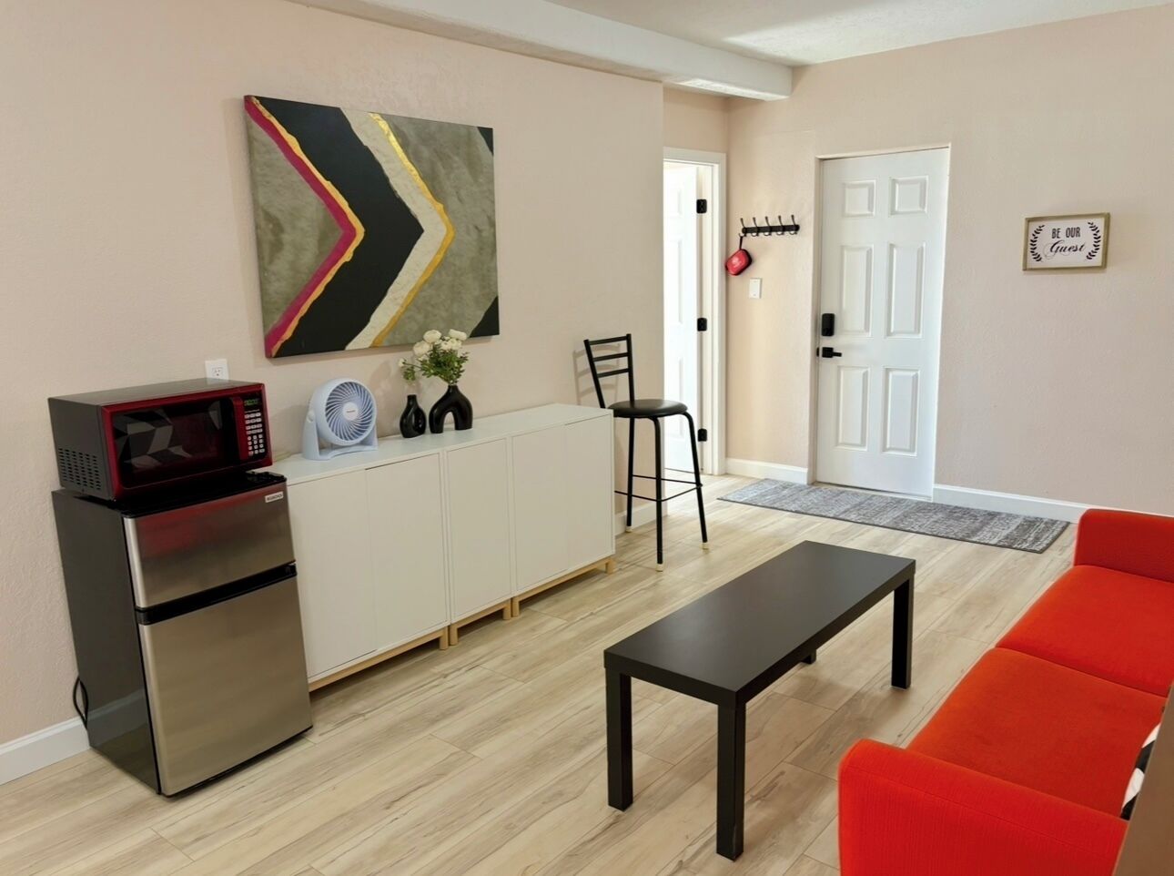 Un espace de vie compact comprenant un coin cuisine, une table à manger, un canapé rouge, une chaise noire, un ventilateur et un élément décoratif mural.