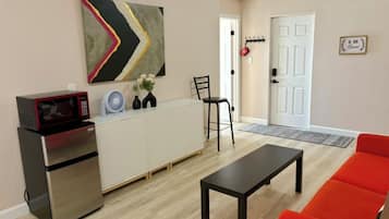 Un espace de vie compact comprenant un coin cuisine, une table à manger, un canapé rouge, une chaise noire, un ventilateur et un élément décoratif mural.