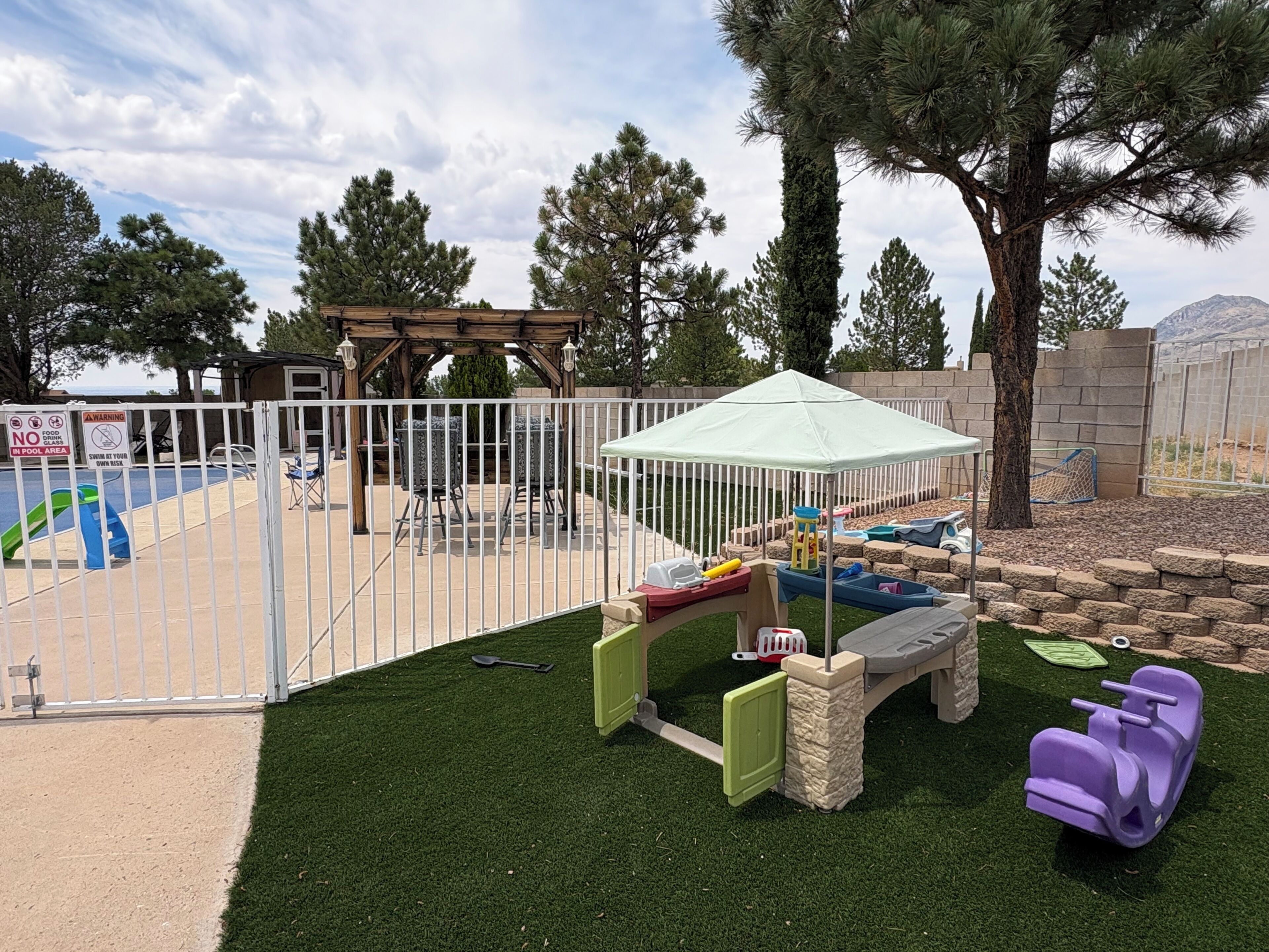 A fenced outdoor area with a pool, a children's play structure, and a gazebo.