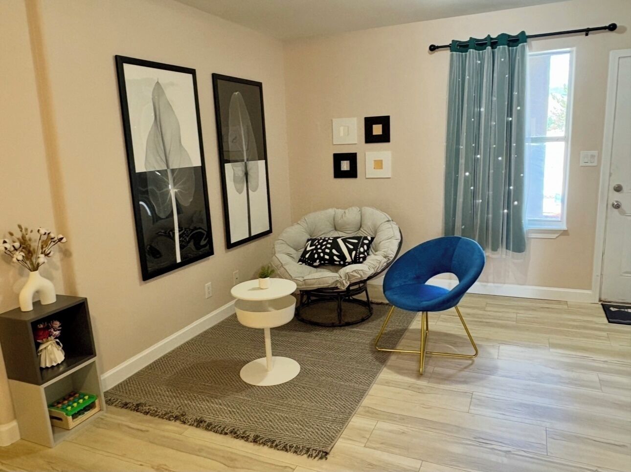 Un salon avec un fauteuil gris, une chaise bleue, une petite table blanche et deux œuvres d’art encadrées accrochées au mur.