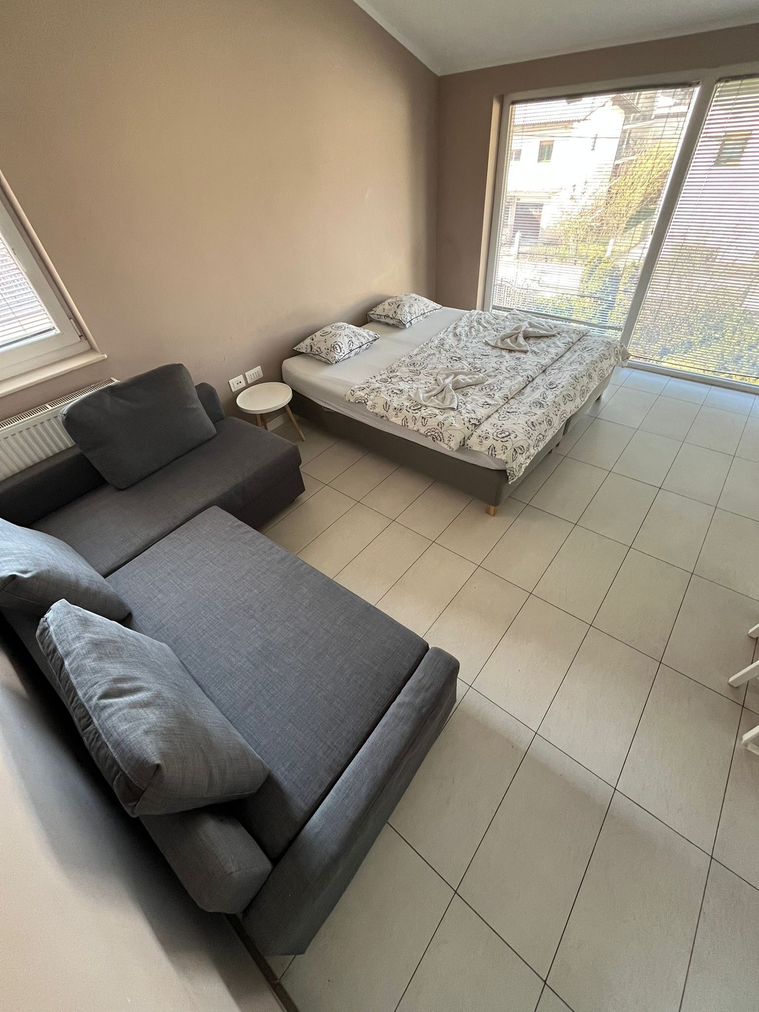 A modern bedroom with a sofa, a small round table, a bed with patterned bedding, and large windows.