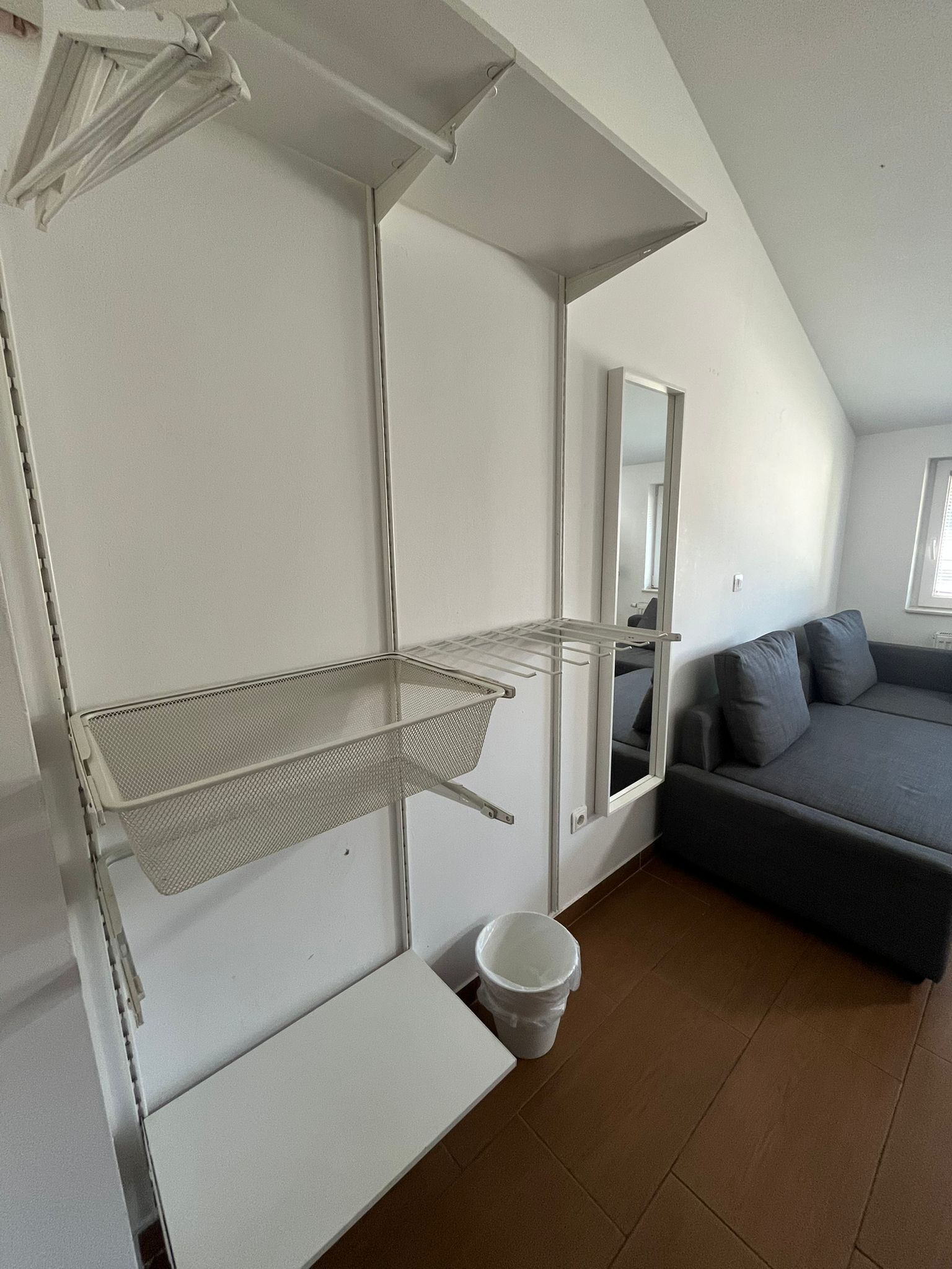 A room with a sloped ceiling, a gray sofa, a white wall-mounted shelf, and a white bucket on the floor.