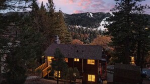 Une cabane aux murs en bois, un escalier menant à l’entrée et, en arrière-plan, une vue sur une chaîne de montagnes boisées.