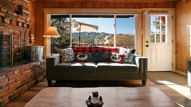 A cozy living room with a fireplace, a sofa with patterned cushions, and a view of a mountainous landscape through large windows. - Cozy Cabin, Mountain Views, Hot Tub, Pool Table (Big Bear Lake)