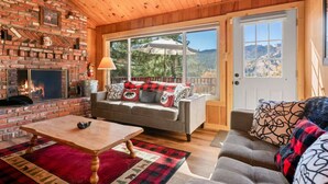 A cozy room with a fireplace, a sofa, a table, and a view of the outdoors. - Cozy Cabin, Mountain Views, Hot Tub, Pool Table (Big Bear Lake)