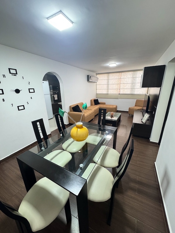 Una sala de estar moderna con comedor, un conjunto de sofás, un televisor y un reloj de pared.
