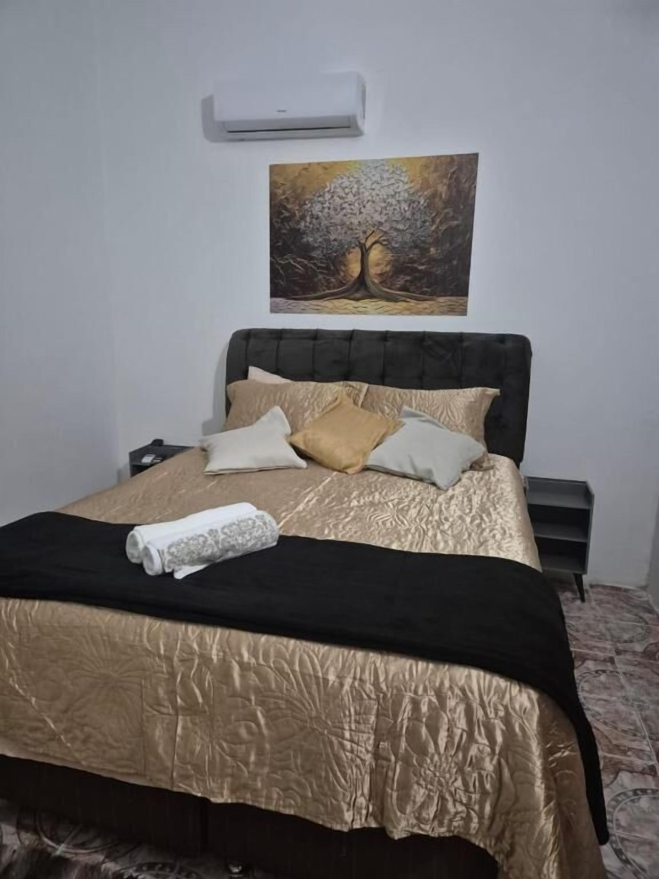 Una cama bien tendida con una colcha dorada y negra, una toalla blanca y un cuadro de un árbol en la pared de arriba.