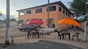 A building with a sandy ground, outdoor seating with umbrellas, and a parked car. - Pousada Imperatriz  (Itaparica)