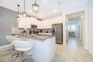 A modern kitchen with white cabinets, a central island with bar stools, stainless steel appliances, and pendant lighting. - Magic Awaits: Modern 3BR Family Retreat (Kissimmee)