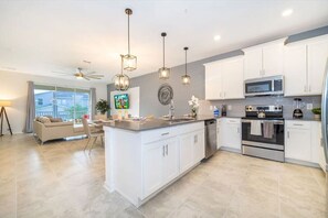 A modern kitchen with white cabinets, a central island, stainless steel appliances, pendant lighting, and a dining area with a sofa and table. - Magic Awaits: Modern 3BR Family Retreat (Kissimmee)
