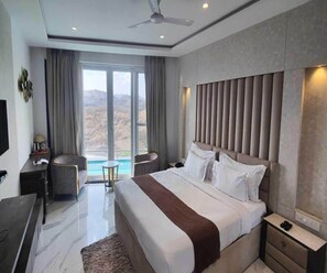 A modern hotel room with a large bed, a desk, a chair, a small table, and a view of the mountains. - Clubhouse Swarnagarh Nature Retreat Udaipur (Udaipur)