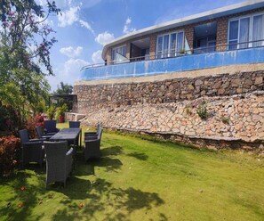 A stone retaining wall, outdoor seating, and a building with large windows. - Clubhouse Swarnagarh Nature Retreat Udaipur (Udaipur)
