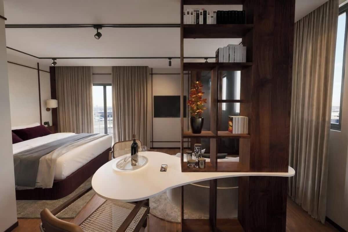 A modern hotel room with a curved desk, a bed, a bookshelf, and a city view.