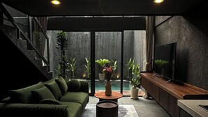 Ein modernes Wohnzimmer mit einem grünen Sofa, einem Couchtisch und einem Fernseher. Pflanzen und eine Treppe sind ebenfalls zu sehen.
