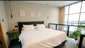 Ein modernes Hotelzimmer mit einem großen Bett, Nachttischen, Blick durch ein Fenster und Kunstwerken an der Wand.