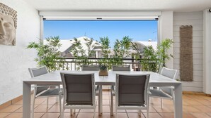 Un patio con mesa y sillas, rodeado de plantas, y vista a edificios al fondo.