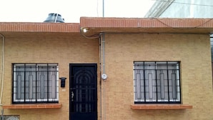 Un edificio de dos pisos con una puerta negra, dos ventanas con barrotes y un cuadro eléctrico visible en la pared.