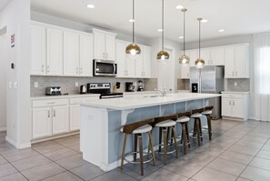 A modern kitchen with white cabinets, a large island with bar stools, stainless steel appliances, and pendant lighting. - Solara 9BR Villa w/ Pool, Spa & Game Room (Kissimmee)