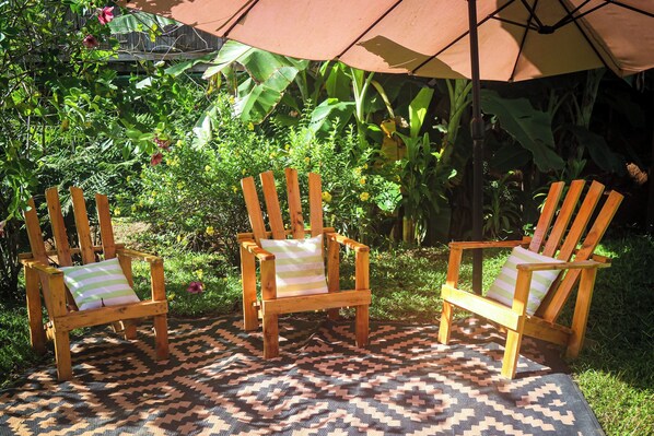 Three wooden chairs with striped cushions, a patterned rug, and an umbrella in a garden setting.