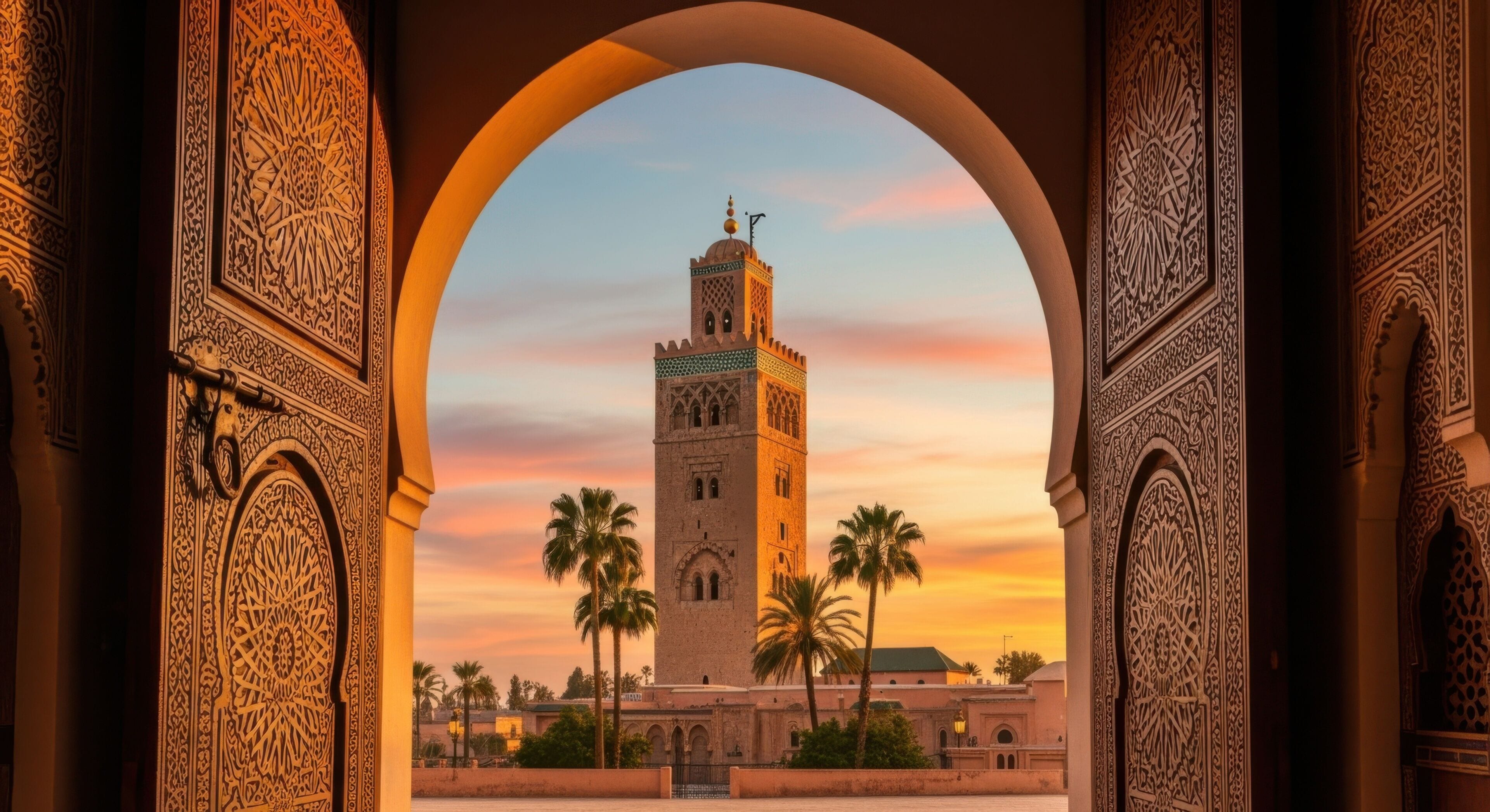 Un arco ornamentado enmarca la vista de una torre histórica con un minarete, palmeras y un cielo al atardecer.