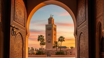 Un arco ornamentado enmarca la vista de una torre histórica con un minarete, palmeras y un cielo al atardecer.
