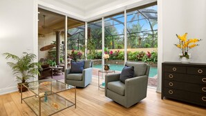 Ein geräumiges Wohnzimmer mit einem Glastisch, zwei grauen Sesseln, einer dunklen Holzkommode und Blick auf einen Swimmingpool durch große Fenster.