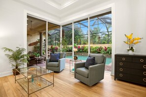 Ein geräumiges Wohnzimmer mit einem Glastisch, zwei grauen Sesseln, einer dunklen Holzkommode und Blick auf einen Swimmingpool durch große Fenster.