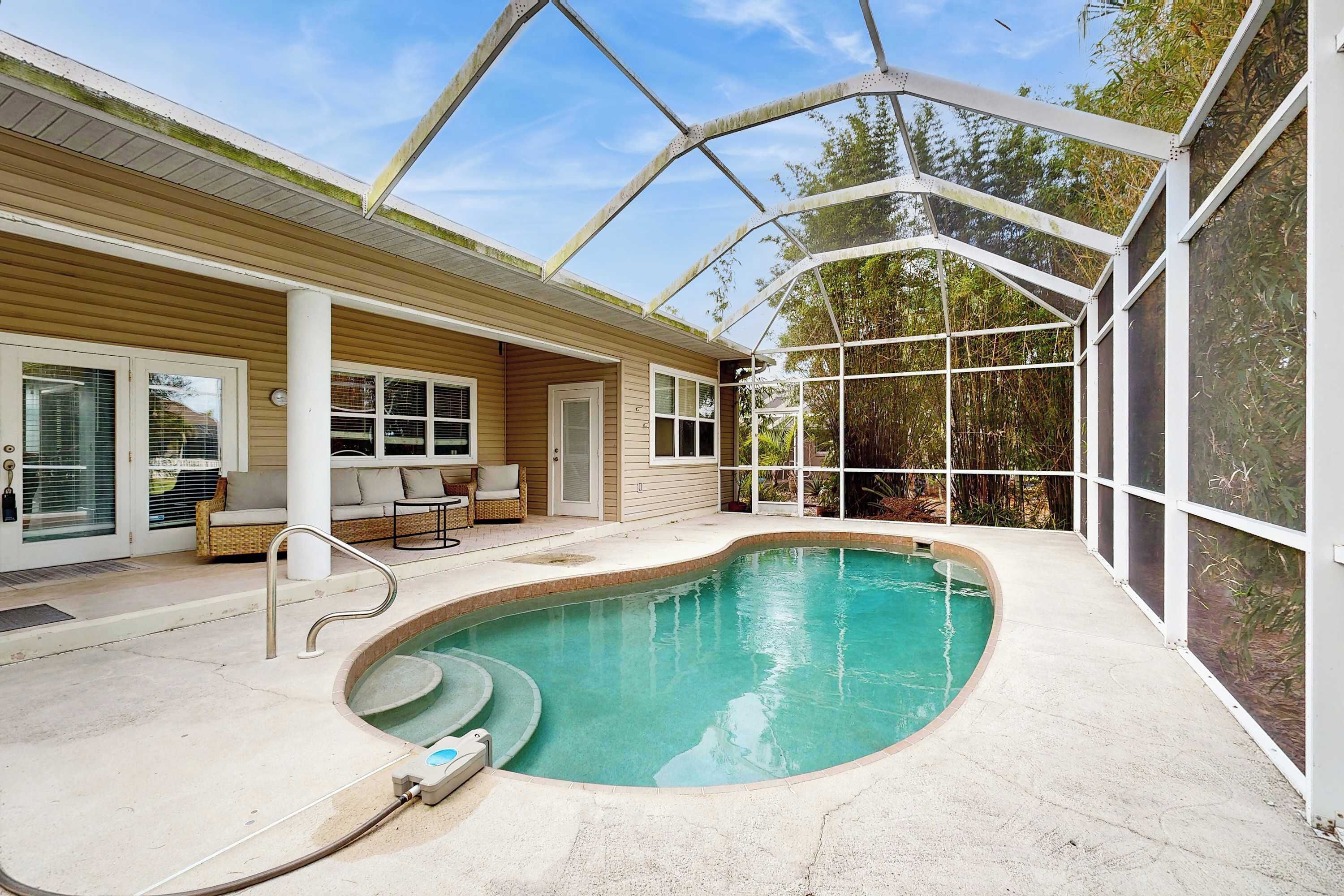 A swimming pool with a glass enclosure, a patio with outdoor furniture, and a covered area with a view of trees.