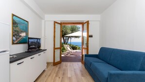 A modern room with a wooden floor, a blue sofa, a kitchen area with a TV, and a view of the sea through an open door. - Apartment 'Paradise Residence 1' with Sea View, Wi-Fi and Air Conditioning (Sant'Alessio Village)