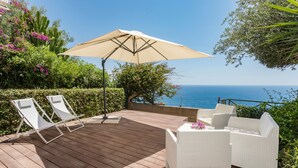 Eine Holzterrasse mit weißen Korbmöbeln, einem Tisch mit einer Blume und Blick aufs Meer.