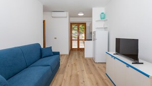 Ein modernes Wohnzimmer mit einem blauen Sofa, einem Flachbildfernseher auf einem weißen Schrank und einer Küchenzeile mit Kühlschrank und einer türkisfarbenen Vase auf einem Regal.