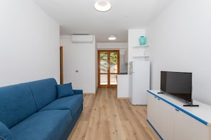 Una sala de estar moderna con un sofá azul, un televisor de pantalla plana sobre un mueble blanco y una zona de cocina con refrigerador y pileta.