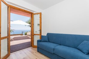 A room with a blue sofa, a wooden door leading to an outdoor deck, and a view of the sea. - Apartment 'Paradise Residence 2' with Sea View, Wi-Fi and Air Conditioning (Sant'Alessio Village)