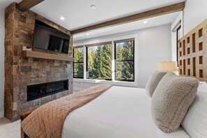 A bedroom with a stone fireplace, a large bed, a TV mounted on the wall, and a window with a view of trees.