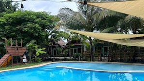A swimming pool with a wooden deck, surrounded by palm trees and a shaded area. - Cabañas Kai  (Iztapa)