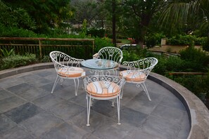 Un patio exterior circular con cuatro sillas blancas de metal y una mesa de vidrio, rodeado de un suelo de piedra y follaje.